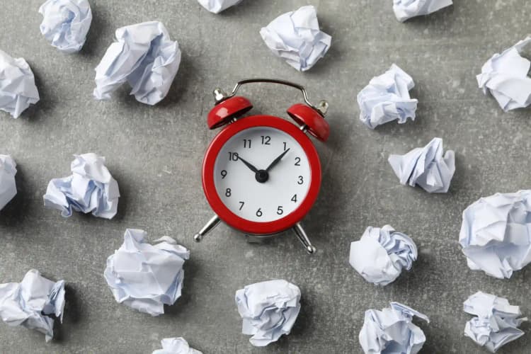 Red alarm clock surrounded by discarded paperwork on a gray desk