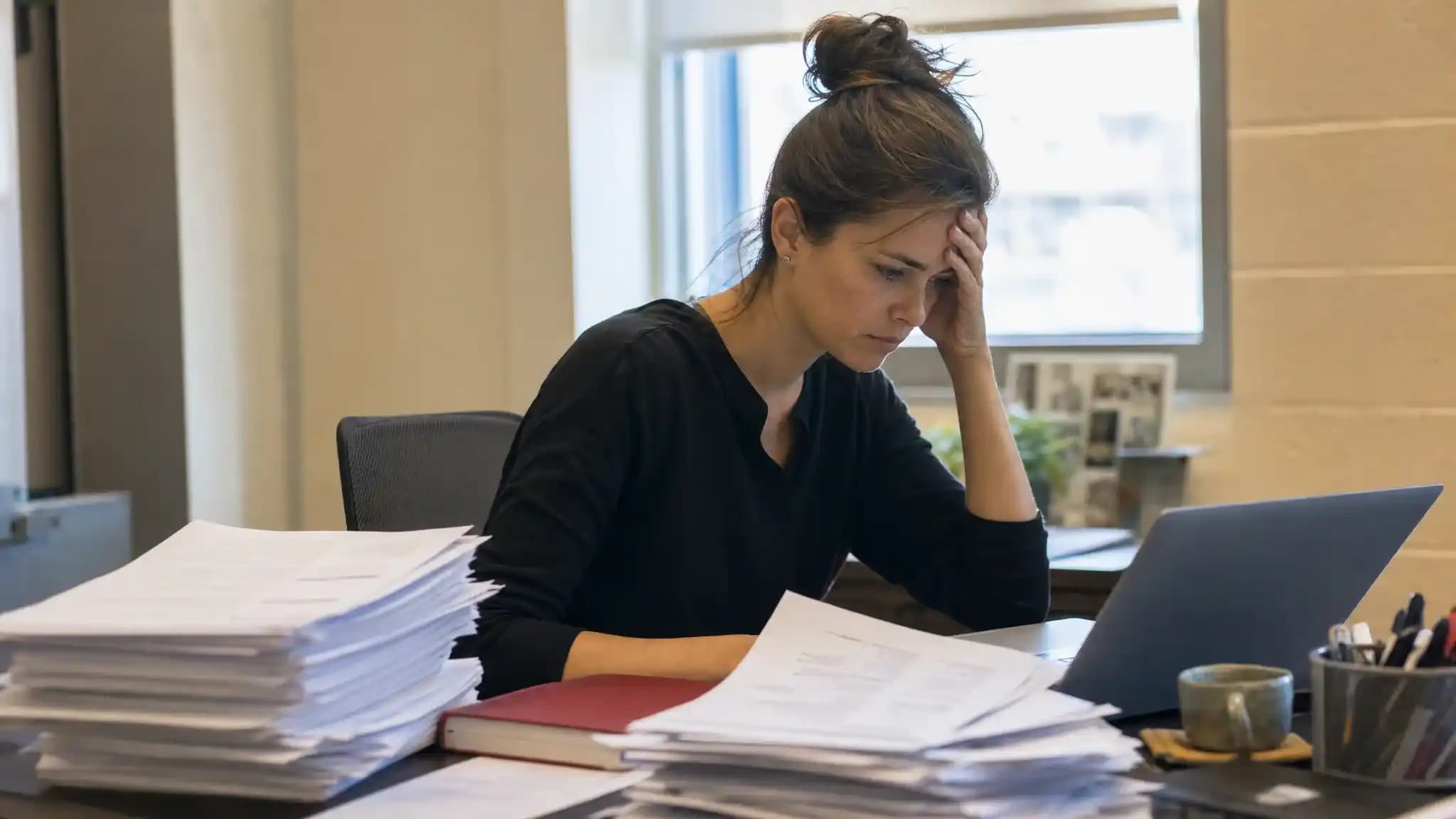 A tired recruiter sits at a desk buried under a tall stack of printed resumes